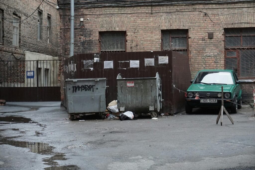 pexels-photo-6455714-6455714 A gritty urban alley featuring garbage bins, scattered litter, and an old parked car.